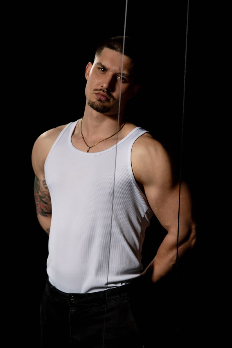 Mann im weissen Tanktop mit Halskette hinter einer Glasscheibe