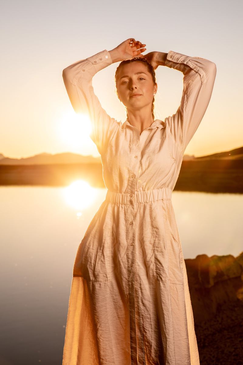 Frau im hellen Kleid hebt die Arme vor einem See bei Sonnenuntergang