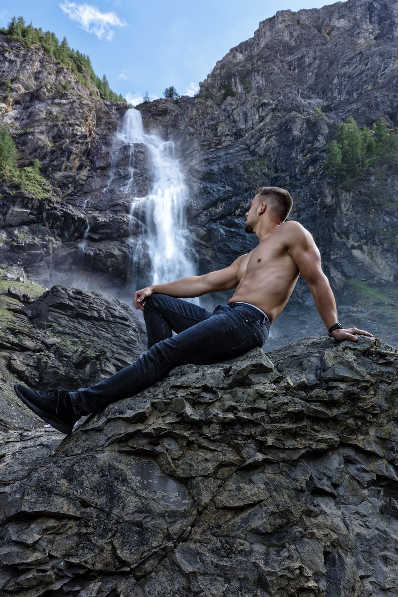 Mann mit freiem Oberkörper auf einem Felsen vor einem Wasserfall